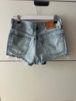 Levi's ljusblå jeansshorts med fransar - Säljer ett par klassiska ljusblå jeansshorts från Levi's med råa fransade kanter och slitningar framtill. Shortsen har fem fickor, bälteshällor och knappgylf. Perfekta för sommaren och ger en avslappnad vibe. Storlek W26, säljer då jag inte använder dom längre 