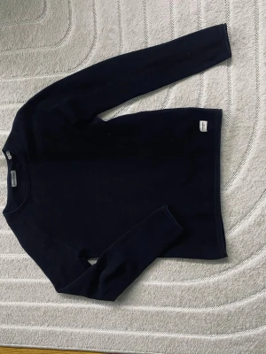 Mörkblå långärmad tröja från Filippa K - En stilren mörkblå långärmad tröja från Filippa K. Tröjan har rund halsringning och en diskret logolapp nertill. Perfekt för en clean och enkel look. Materialet känns mjukt och följsamt mot kroppen.