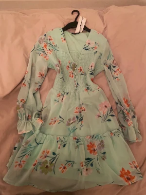 Mintgrön blommig klänning med volang - Superfin klänning med blommigt mönster. Klänningen har långärmade ärmar med volang vid ärmslut, v-ringning och smockad midja. Luftigt material, perfekt för varmare dagar.