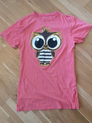 Korall/Rosa t-shirt med uggletryck - Söt rosa t-shirt med stort tryck av en tecknad uggla med stora ögon och randig mage på framsidan. T-shirten är även väldigt lång och passar perfekt som sovplagg