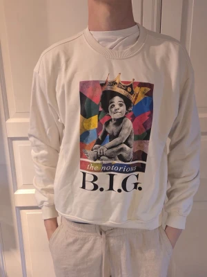 Vit sweatshirt The Notorious B.I.G. - Vit sweatshirt med färgglatt tryck av The Notorious B.I.G. på framsidan. Tröjan har rund halsringning, ribbade muddar och lång ärm. Perfekt för dig som gillar streetwear och musikikoner.