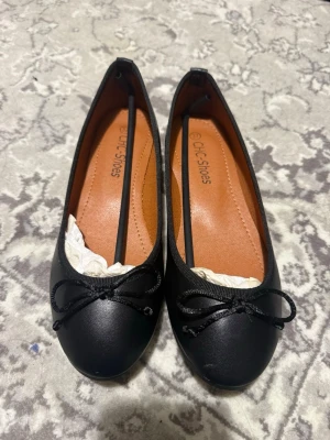 Svarta ballerinaskor från CLC Shoes - Stilrena svarta ballerinaskor från CLC Shoes med rund tå och dekorativ rosett framtill. Skorna har en slät ovandel i syntetmaterial och en bekväm innersula i brunt. Perfekta för en klassisk och enkel look.