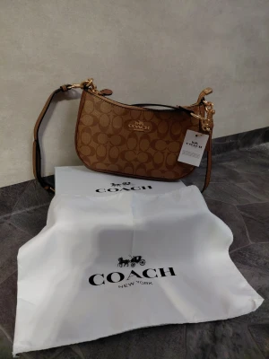 Brun handväska från Coach - Snygg brun handväska från Coach med klassiskt monogrammönster och guldfärgade detaljer. Väskan har justerbar axelrem och levereras med original dustbag. Perfekt för dig som gillar stilrena accessoarer med lyxig känsla.