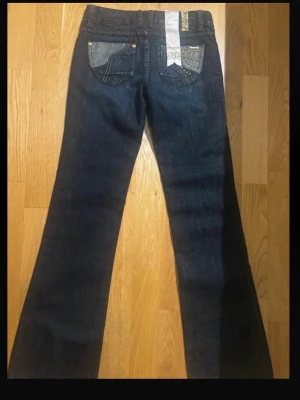 Mörkblå vintage jenas  - Snygga mörkblå bootcut jeans  med klassiska fem fickor och dekorativa detaljer på bakfickorna. Jeansen har låg midja och är tillverkade i denim med en liten stretch för skön passform. Perfekta för en trendig och avslappnad stil.