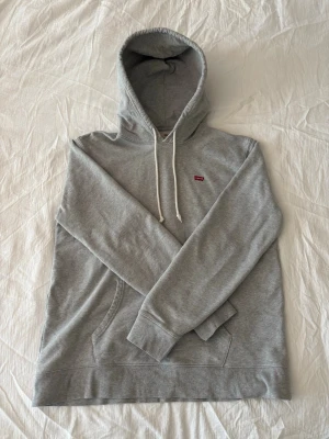 Grå hoodie från Levi's - Snygg grå hoodie från Levi's med klassisk röd logga på bröstet. Känguruficka och ribbade muddar vid ärmslut och nederkant. Perfekt för en avslappnad och stilren look.