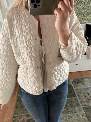 Vit vårjacka från Zara - Superfin vit quiltad cardiganjacka från Zara med lätt boxig passform och tryckknappar framtill. Jackan har ett diskret mönster i tyget och är perfekt att slänga på över en topp. Lätt vadderad och med långa ärmar.