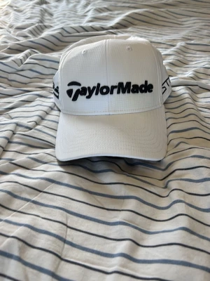 Vit TaylorMade keps med brodyr - Snygg vit keps från TaylorMade med svart broderad logga framtill och TP5-tryck på sidan. Kepsen har böjd skärm och justerbar passform. Tillverkad i lätt och ventilerande material, perfekt för golf eller streetwear.