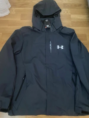 Svart vindjacka från Under Armour - Svart vindjacka från Under Armour med huva, dragkedja och två sidofickor. Jackan har vitt logotryck på bröstet och texten 'Explore' längs dragkedjan. Tillverkad i lätt och vattenavvisande material, perfekt för blåsiga dagar.