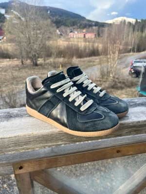Svarta Maison margielas  - Snygga svarta sneakers med kontrasterande vita skosnören och beige innersula. Skorna har ovandel i både mocka och läder, samt en klassisk gummisula i ljusbrunt. Perfekta för dig som gillar stilrena och tidlösa sneakers med retrovibbar. ! Dom är lite trasiga i hälen och lite däf fram men det är inget man tänker på 