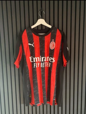 AC Milan matchtröja Puma röd/svart - Säljer en AC Milan matchtröja från Puma i klassiska röda och svarta vertikala ränder. Tröjan har klubbmärke och Puma-logga på bröstet samt Emirates Fly Better-tryck på magen. Korta ärmar och rund hals. Perfekt för fotbollsfans!