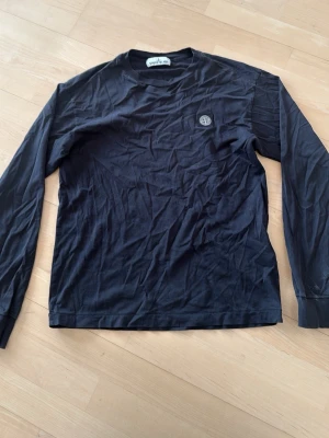 Svart långärmad tröja från Stone Island - Svart långärmad tröja från Stone Island med rund hals och diskret logomärke på bröstet. Tillverkad i mjuk bomull som känns skön mot huden. Perfekt för en stilren och avslappnad look.