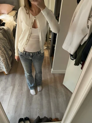 Vit kofta från Holly&whyte - Snygg vit kofta från Holly&whyte med rund halsringning och knappar framtill. Långärmad modell i mjukt, tunt material som passar perfekt till lager på lager. Diskret logga och ribbade muddar vid ärmslut och nederkant.