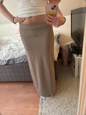 Beige långkjol med slits - Stilren beige långkjol med slits bak. Kjolen har en rak passform och är tillverkad i ett mjukt och stretchigt material som sitter snyggt på kroppen. Perfekt att matcha med en crop top eller stickad tröja.