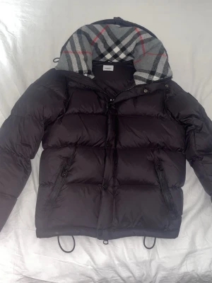 burberry jacka - Svart pufferjacka från Burberry med klassisk rutmönstrad huva i grått, vitt och rött. Jackan har dragkedja framtill, två fickor med dragkedja och justerbar dragsko nedtill. Perfekt för kalla vinterdagar och riktigt snygg streetstil.