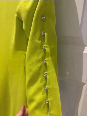 Gul klänning från Karen Millen - Snygg gul klänning från Karen Millen med v-ringning och cut-out detalj framtill. Klänningen har långa ärmar med dekorativa knappar längs ärmslutet och en figurnära passform. Perfekt för dig som vill sticka ut med färg och stil.