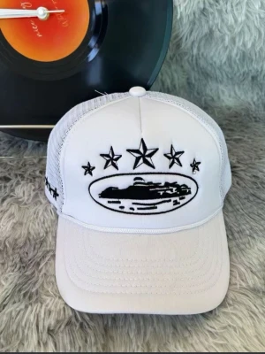 Vit meshkeps med broderade stjärnor - Snygg vit keps med mesh på sidorna och broderade svarta stjärnor samt motiv framtill. Justerbar baktill för perfekt passform. Perfekt accessoar för en streetwear-look.
