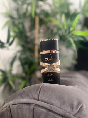 Lattafa Asad parfym svart/guld 100ml - Lattafa Asad parfymflaska i matt svart med eleganta gulddetaljer och arabiska bokstäver. Flaskan har en unik form med guldband runt och ett dekorativt emblem framtill. Perfekt för dig som gillar exklusiv design och orientaliska dofter.