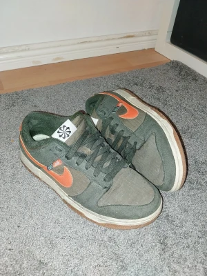 Nike Dunk Low  - Nike Dunk Low sneakers i mörkgrönt tyg och mocka med orange swoosh och hälpanel. Vit mellansula och brun yttersula. Klassisk låg modell med snörning och Nike-logga på hälen. Perfekta för dig som gillar streetwear och vill sticka ut med färg skorna är även fodrade och storlek 40.5 perfekt för dig som skatear då det var då jag själv använde dom här skorna.