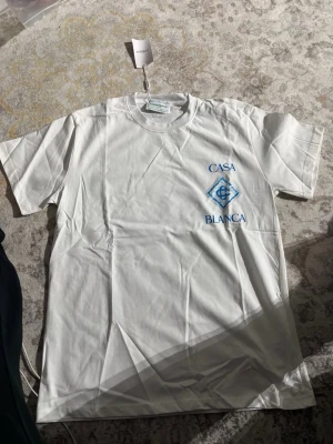 Vit Casa Blanca t-shirt med blått tryck - Vit t-shirt från Casa Blanca med blått tryck och logga både fram och bak. Klassisk passform med rund hals och korta ärmar. Tillverkad i mjuk bomull, perfekt för en clean och stilren look.