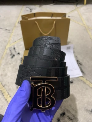 Burberry bälte med TB-spänne - Säljer 2 sidig Burberry bälten i skinn med klassiskt TB-spänne i metall. En sida är svart med diskret rutmönster och andra sidan är beige med brunt rutmönster. Båda sidor har präglad Burberry-logga och känns riktigt lyxigt. Perfekt accessoar för att lyfta din outfit.