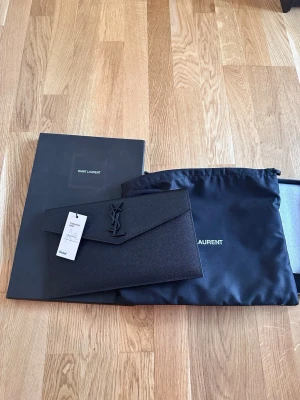 Svart kuvertväska från Saint Laurent - Snygg svart kuvertväska från Saint Laurent i strukturerat läder med ikoniska YSL-loggan framtill. Stilren och elegant design med magnetisk stängning. Kommer med originalbox och dustbag.