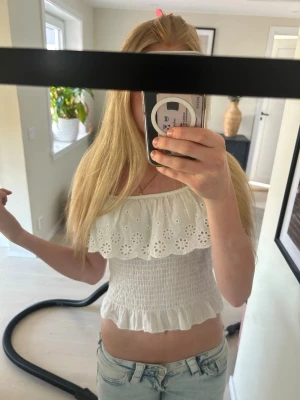 Vit offshoulder topp med volang - Superfin vit offshoulder topp med broderad volang och smockad midja. Toppen är ärmlös och har en söt, romantisk känsla med volangdetaljer både upptill och nertill. Perfekt till jeans eller kjol för en fräsch look.
