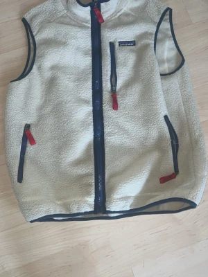 Beige fleeceväst från Patagonia - Säljer en beige fleeceväst från Patagonia med blå dragkedjor och röda detaljer. Västen har tre fickor med dragkedja och är kantad med blått tyg. Perfekt för lager-på-lager och outdoor-stil.