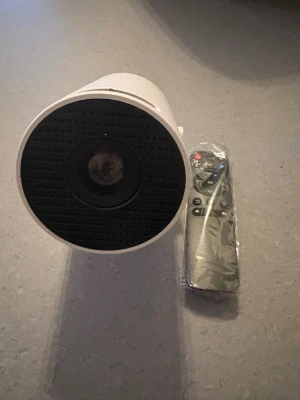Vit projektor med fjärrkontroll - Säljer en vit projektor med rund lins och svart front. Medföljer en smidig fjärrkontroll som fortfarande är inplastad. Perfekt för filmkvällar eller gaming. Projektorn har en modern design och är tillverkad i plast.