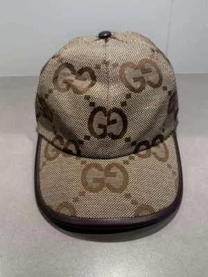 Beige keps från Guccic med GG-mönster - Snygg beige keps från Guccic med klassiskt GG-monogram i brunt över hela kepsen. Kepsen har böjd skärm med mörkbrun kant och är tillverkad i canvasmaterial. Perfekt accessoar för en trendig streetstyle-look.