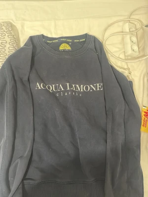 Mörkblå sweatshirt Acqua Limone - Mörkblå sweatshirt från Acqua Limone med vit logotyp framtill och stor gul citronlogga på ryggen. Klassisk rund halsringning och ribbade muddar. Perfekt för en avslappnad och trendig stil.