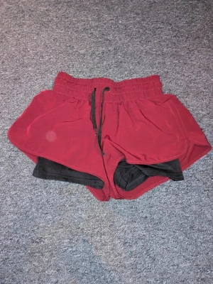 Vinröda träningsshorts med innershorts - Vinröda träningsshorts med svarta innershorts och snörning i midjan. Shortsen har elastisk midja och är tillverkade i ett syntetmaterial som är lätt och snabbtorkande. Perfekta för träning och löpning, med sportig och bekväm passform.