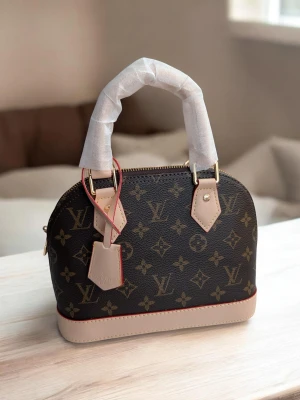 Iouis Vuitton Alma BB  - En elegant handväska i klassiskt Monogram-canvas med detaljer i ljust vachetta-läder. Väskan har den ikoniska strukturerade kupolformen och är utrustad med dubbla handtag samt en matchande clochette (nyckelhållare).