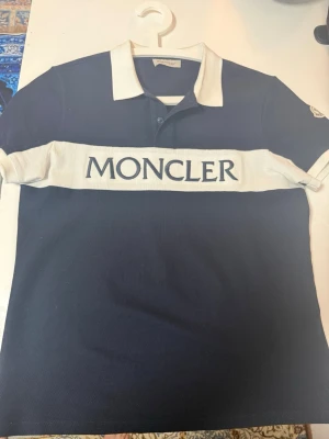 Moncler pikétröja med logga - Mörkblå pikétröja från Moncler med vit krage och bred vit rand över bröstet där Moncler-loggan är broderad. Klassisk pikédesign med knappar vid halsen och korta ärmar. Snygg och stilren, perfekt för en trendig look.