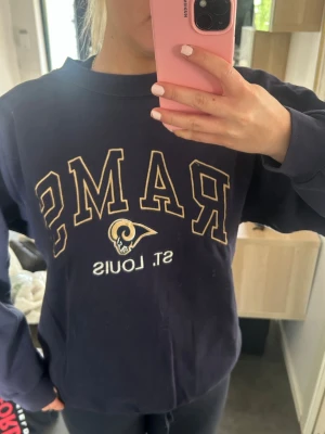 Blå St. Louis Rams sweatshirt - Mörkblå sweatshirt med St. Louis Rams-logga och text framtill. Tröjan har rund halsringning, långa ärmar och ribbade muddar. 