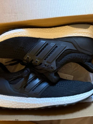 Adidas Ultraboost svarta sneakers - Svarta Adidas Ultraboost sneakers med stickad ovandel och klassiska tre ränder på sidan. Skorna har en vit, responsiv Boost-sula och svart yttersula med grepp. Perfekta för dig som gillar sportig och modern stil. Snygg design och bekväm passform.