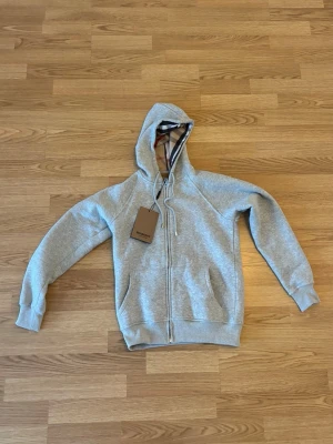 Grå zip-hoodie från Burberry - Snygg grå hoodie med dragkedja från Burberry. Klassisk huva med det ikoniska rutmönstret på insidan, dragsko och två fickor framtill. Mjuk bomullskänsla och ribbade muddar vid ärmslut och midja. Perfekt för en chill och stilren look. OBS INTE ORG1NAL