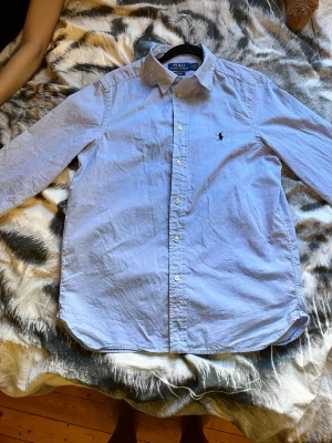 Ljusblå skjorta från Polo Ralph Lauren - Klassisk ljusblå skjorta från Polo Ralph Lauren med button-down krage och broderad logga på bröstet. Skjortan har långa ärmar och knäppning framtill. Perfekt för en stilren och avslappnad look.