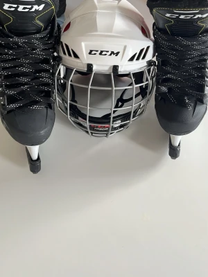 CCM hockeyhjälm och skridskor - Säljer ett set med svarta CCM hockeyskridskor och en vit CCM hockeyhjälm med galler. Skridskorna har svarta och vita snören och robust konstruktion, hjälmen har justerbart galler för extra skydd. Perfekt för hockeyträning eller match. Storlek på hjälmen är S och skridskorna är 40,5