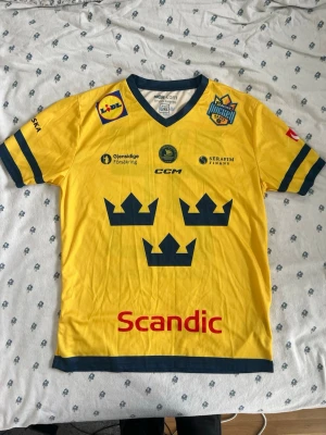 Gul Tre Kronor hockeytröja CCM - Gul hockeytröja från CCM med Tre Kronor-tryck på bröstet och nummer 25 på ryggen. Tröjan har blå detaljer vid ärmslut, krage och nederkant  nosamt flera sponsormärken. Perfekt för dig som vill stötta svenska landslaget.