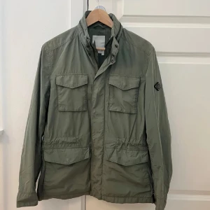 Grön fieldjacket från J.Lindeberg - Snygg grön jacka från J.Lindeberg med dragkedja och knappar. Den har flera praktiska fickor och en stilren design. Perfekt för höst och vår. Priset ej hugget i sten litet hål som jag märkte nu när jag ska sälja den finns men man märken den knappt                Nypris : Runt 3500