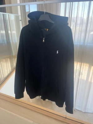 Svart hoodie från Polo Ralph Lauren - Svart hoodie med dragkedja från Polo Ralph Lauren. Klassisk design med huva, fickor framtill och den ikoniska vita loggan broderad på bröstet. Tillverkad i mjukt bomullsmaterial som är skönt att bära. Perfekt för en avslappnad och stilren look. Det är en XL i barn så det är som en vuxen M men den sitter lite mer som en S