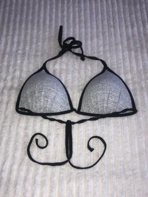 Svart och grå trekantsbikini - Snygg trekantsbikini med grå kupor och svarta kanter. Klassisk modell med knyt i nacken och ryggen, perfekt för stranden eller poolen. Enkel och stilren design som passar till det mesta.