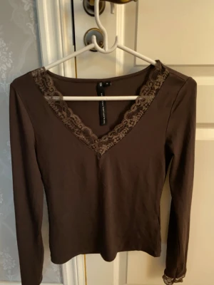 Brun långärmad topp med spets - Snygg brun långärmad topp från lager 157 med  V-ringning och dekorativ spetskant runt halsen. Mjuk och stretchig bomullsblandning som sitter skönt på kroppen. Använd en gång. 