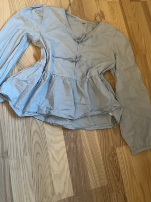 Ljusblå blus från Gina Tricot - Supersöt ljusblå blus från Gina Tricot med v-ringning och volang nedtill. Blusen har långa ärmar och knappar framtill. Perfekt att styla med jeans eller kjol för en trendig look.