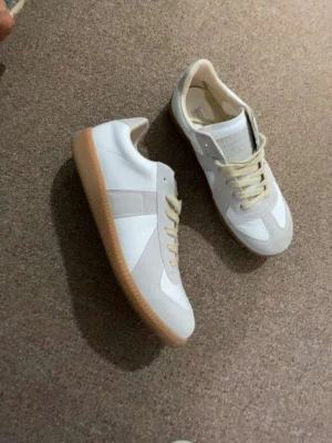 Vita och beige sneakers med gummisula - Snygga sneakers i vitt och beige med klassisk design. Ovandelen är i en mix av mocka och slätt läder, och skorna har beige snören samt en retro-inspirerad gummisula. Perfekta för dig som gillar stilrena och tidlösa sneakers.