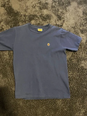 Blå BAPE t-shirt med liten logga - Snygg blå t-shirt från BAPE med klassisk rund hals och liten broderad logga på bröstet. Tillverkad i mjuk bomull och har en enkel, clean design som passar perfekt till streetwear-stilen.