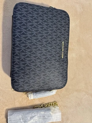 Svart Michael Kors handväska - Snygg svart handväska från Michael Kors med guldfärgad logga och kedjedetaljer. Väskan har ett diskret mönster med MK-loggor över hela ytan och är tillverkad i tåligt konstläder. Perfekt storlek för det viktigaste och en stilren accessoar till din outfit.