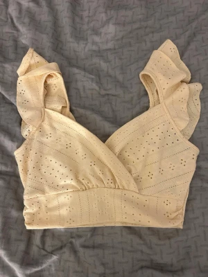 Gul omlottopp med volangärmar - Söt gul omlottopp med volangärmar och broderade hål i tyget. Toppen har en omlott-design och är ärmlös med breda axelband och volangdetaljer. Köpt från SHEIN i storlek xs, använd ett fåtal gånger 