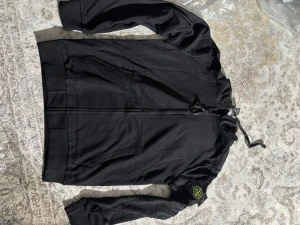 Svart hoodie från Stone Island - Svart hoodie från Stone Island med dragkedja framtill och klassisk logopatch på vänster ärm. Hoodien har två fickor och ribbade muddar. Perfekt för dig som gillar streetwear och vill ha en stilren look.