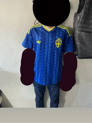 Blå Sverige fotbollströja Adidas - Säljer en blå fotbollströja från Adidas med Sveriges landslagsemblem på bröstet. Tröjan har gula detaljer, korta ärmar och ett mönster i olika blå nyanser. Perfekt för dig som vill visa ditt stöd för Sverige på matchen! Strolek M